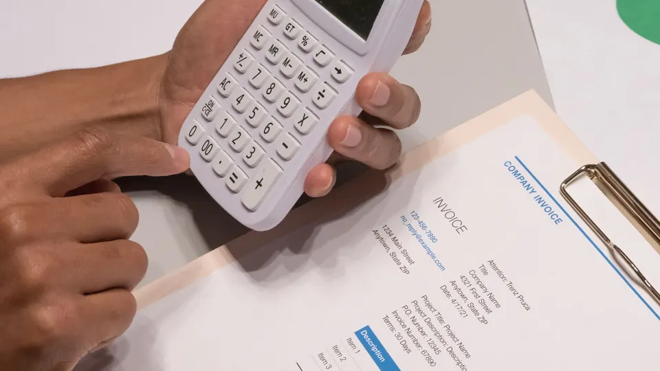 Hands using a calculator next to a printed invoice on a clipboard