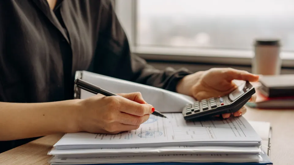 A person writing on paperwork with a calculator on a desk