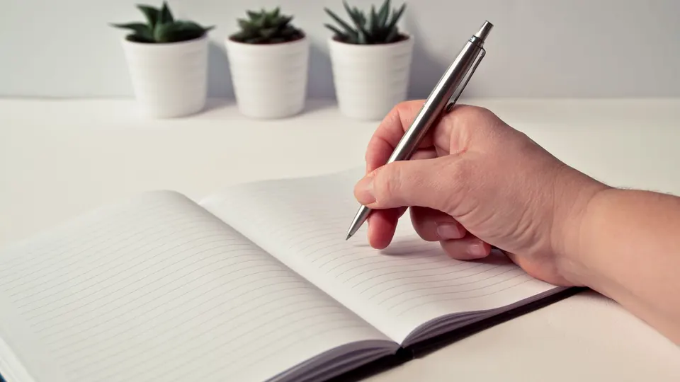 A hand writing in an open lined notebook with a pen on a light desk