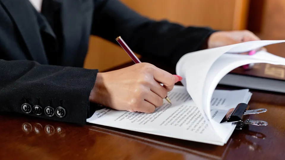 A hand writing with a pen on printed paperwork at an office desk
